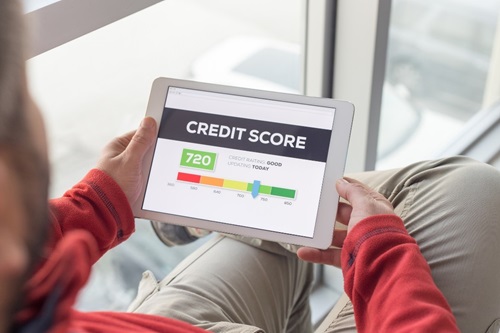 An over-the-shoulder view of a man holding a tablet in his lap. He wears beige pants, a red long sleeve shirt but is otherwise unidentifiable. The tablet shows the bold headline "Credit score" and illustrates a credit score of 720 along a rainbow-hued scale of scores (red, orange, yellow, light green, green). 720 is shown on the light green section of the scale. "Credit rating: good" is also displayed.