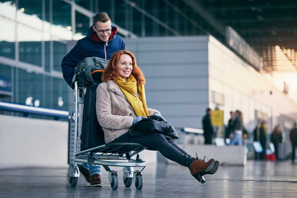 Photo of a scene outside an airport. A man is pushing a redheaded woman on a baggage cart along with their bags. They are both smiling, she has her purse in her lap and her feet lifted up.